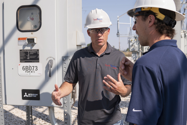Two men in hardhats talk inside a substation